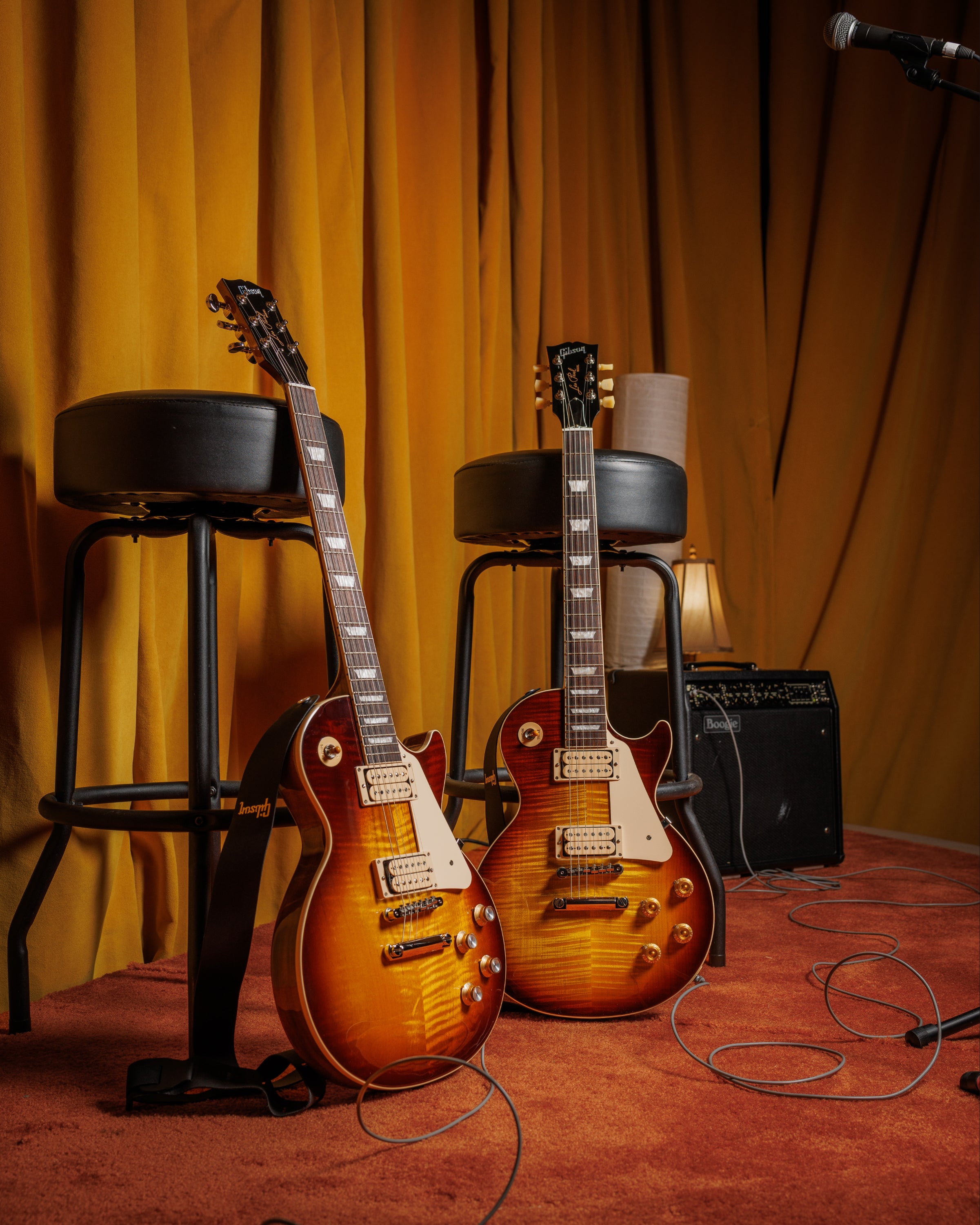 Two electric guitars on stools with a microphone and amplifier in a warm-toned room.
