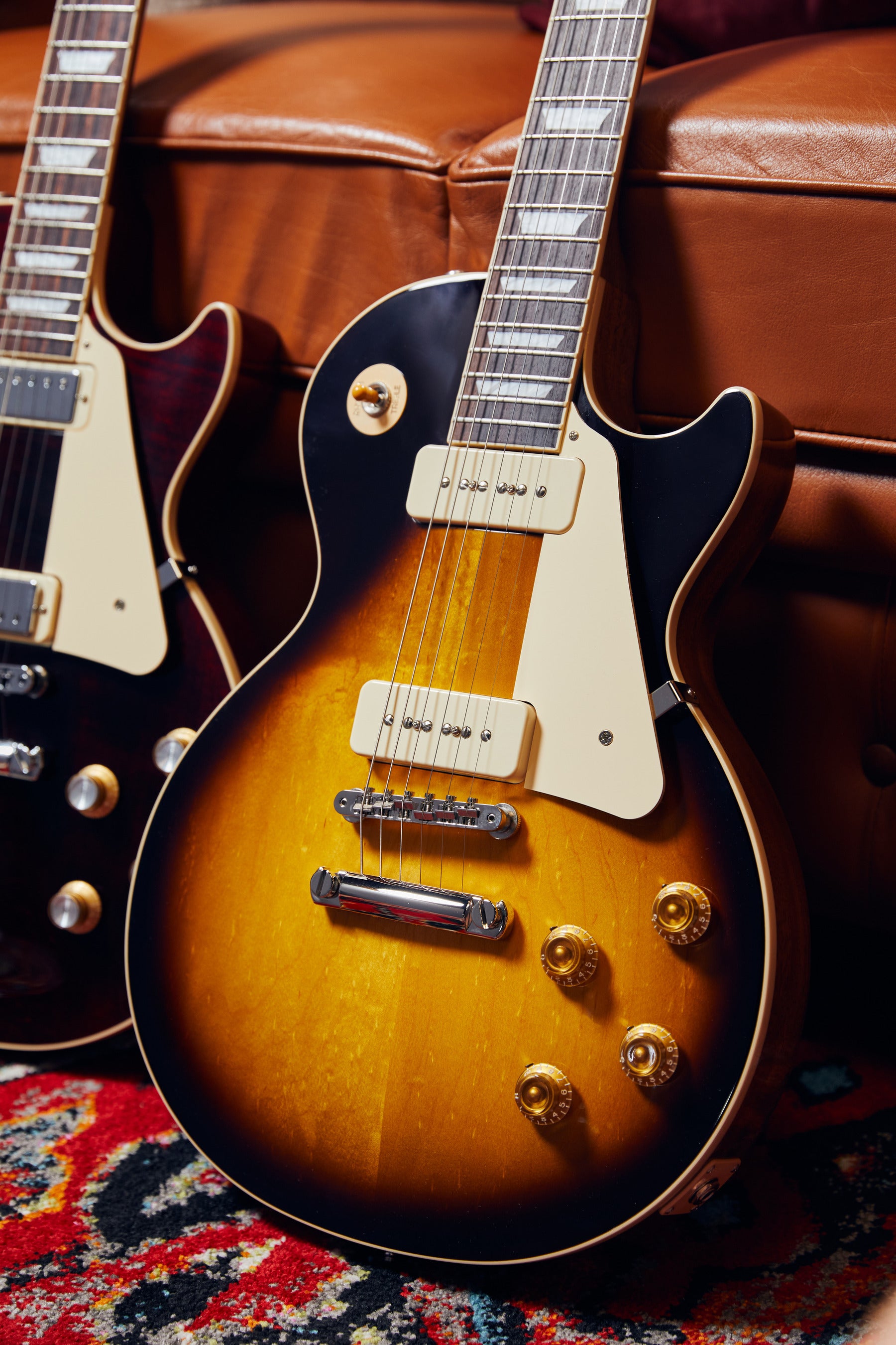 Two electric guitars on a brown leather couch with a colorful rug underneath.